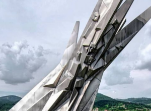ALUMINIJUMSKI MONOLIT U ČAČKU  Visok 10 m i dugačak 17 m, koji, čini se, izbija iz zemlje, sa hiperstilizovanim ljudskim licima